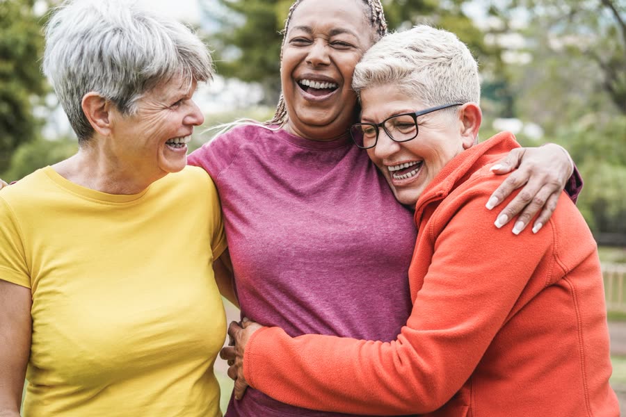 Three older women laughing and hugging outdoors