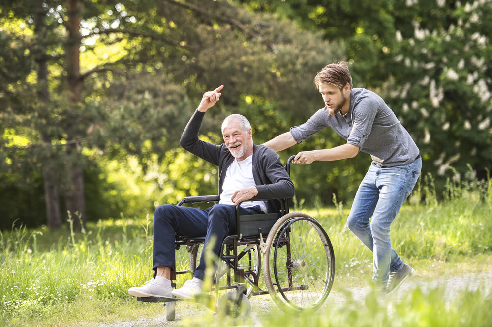 A young man playfully pushing his smiling father's wheelchair through a park